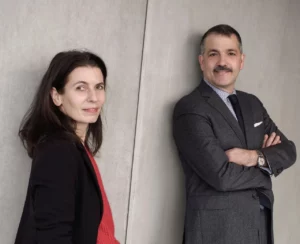 Two professionals pose against a light concrete wall: woman in a red top and black blazer on the left, man in a gray suit with a striped shirt on the right, arms crossed.