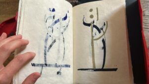 Open sketchbook showing two minimalist abstract ink figures, one on each page; a hand rests on the left edge.