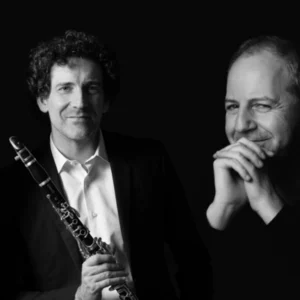 Two male musicians in black-and-white, one holding a clarinet, posed against a dark background.