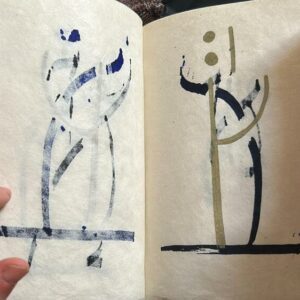 Open notebook showing two pages of abstract ink drawings minimalist humanoid shapes with vertical bodies and circular heads on a cream background