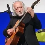 Older man with white hair and beard playing an acoustic guitar on stage wearing a black blazer and concentrating on his music stills a microphone nearby