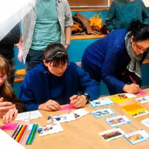 People gathered around a table arranging photos and colored notes for an anti-Semitism awareness mural, with a large geometric banner and logos in the background.