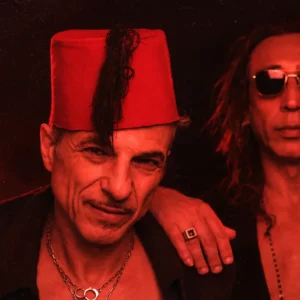 Two men pose together in red lighting; the man on the left wears a red fez and necklace, the man on the right wears sunglasses and long hair.