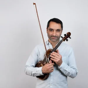 Man in a light blue shirt holding a violin and bow, posing against a plain background.