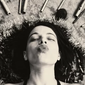 Woman lying with eyes closed, lips puckered to kiss, encircled by beads, tinsel, and craft supplies arranged in a circular frame.
