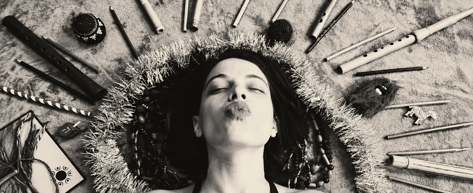 Woman lying with eyes closed lips puckered to kiss encircled by beads tinsel and craft supplies arranged in a circular frame