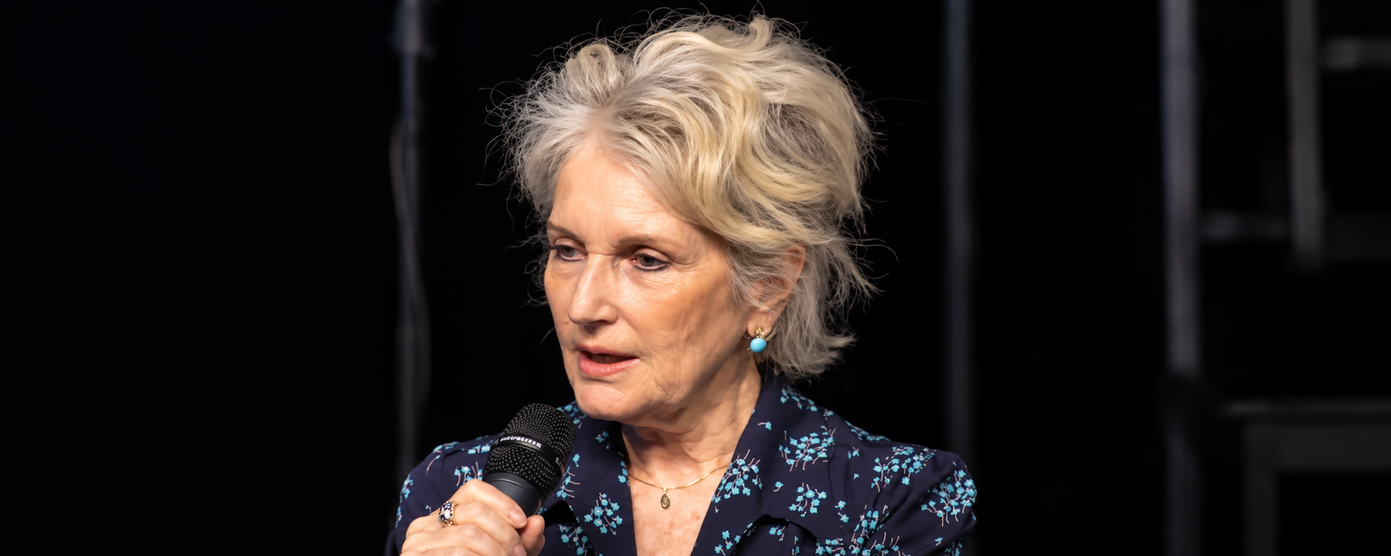 Older woman with short gray hair speaks into a handheld microphone on a dark stage wearing a navy floral blouse