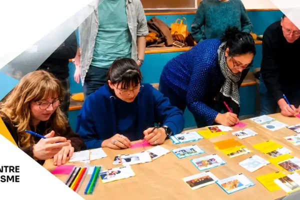 People gathered around a table arranging photos and colored notes for an anti-Semitism awareness mural, with a large geometric banner and logos in the background.