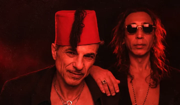 Two men pose together in red lighting; the man on the left wears a red fez and necklace, the man on the right wears sunglasses and long hair.