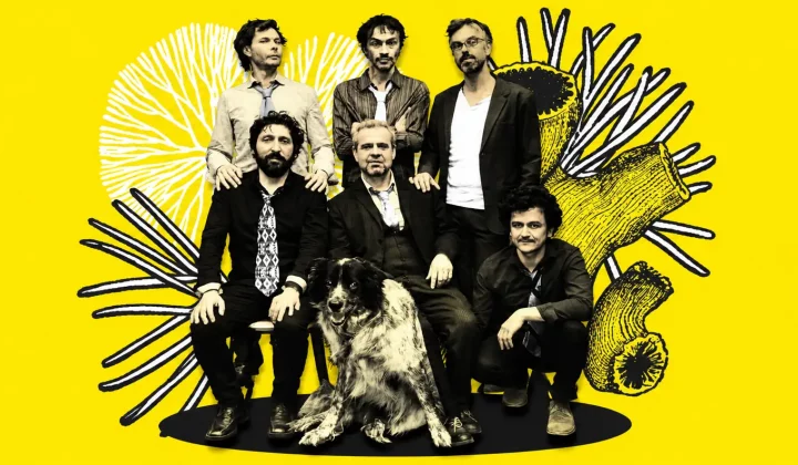 Band photo: six men with a large dog posed on a black platform against a bright yellow backdrop with black abstract drawings.